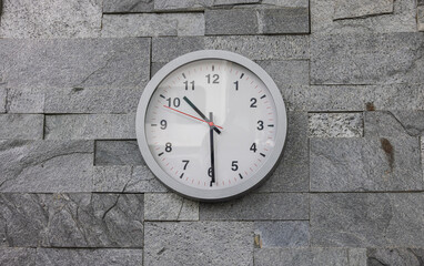 Modern wall clock on a wall tiled with rustic tiles. Modern white wall clock in sunlight with shadows on clock face on a grey background. Close up to a clock, with a red second pointer