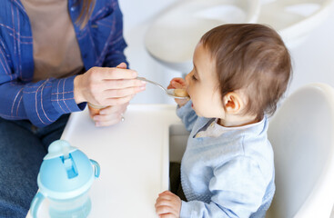 cropped mother feed little toddler baby boy with baby food, from plastic spoon in high chair front view. Motherhood concept. Kitchen lifestyle.Cpose up
