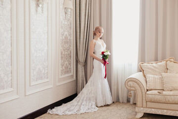 Pretty young Bride in elegant wedding dress with her bouquet. Blonde-haired woman with wedding hair-style in royal room of hotel. Boudoir morning of the bride in luxury interior. Wedding day ceremony