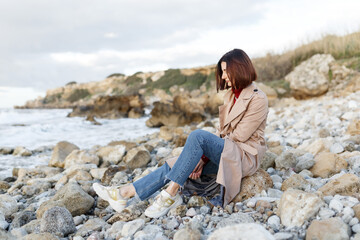 Naklejka na ściany i meble A beautiful young woman sits on a rocky shore and thinks about something. Evening portrait of a beautiful young woman in nature. Solitude, retreat, isoloation, seclusion concept