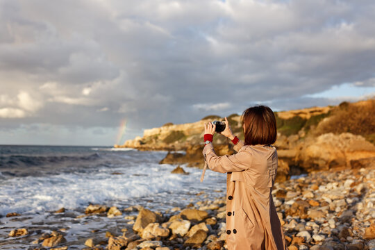 Candid photo of hipster girl traveler on stone shore at winter wearing coat. Fashionable young woman with short red hair makes photo of ocean using camera.