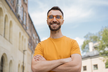 Portrait of handsome smiling man looking at camera, standing on the street. Young confident businessman with arms crossed wearing stylish eyeglasses posing for pictures outdoors