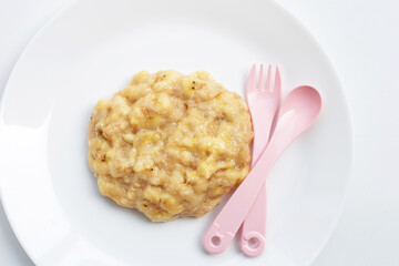 Mashed banana fruit in white plate with pink spoon and fork. Baby food