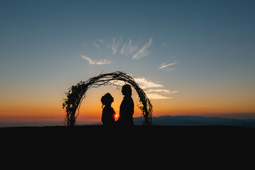 Silhouettes of bride and groom holding hands near the wedding arch at sunset at the wedding ceremony