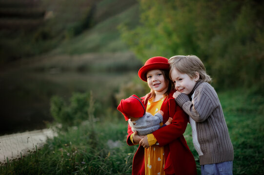 Cute Caucasian girl in red coat and hat with boy playing family with doll baby and puppet stroller in park, children like parents with child hugging, game concept and parenthood, family values