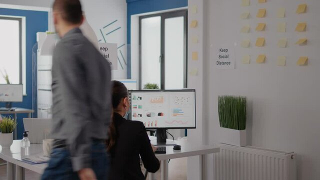 Caucasian male with protective face mask to avoid coronavirus, touching elbow with his collegue while working on technology project sitting at table desk in business office during covid19 lockdown