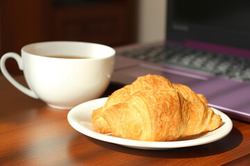 White cup with black tea and croissants in a saucer on the work table. Remote work at home. Snack near the laptop. Sweet morning.