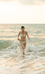 Woman in white bikini walking on sand beach with sunset. Female in swimwear walk on the sand on big waves.