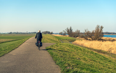Obraz premium An unidentified elderly man with gray hair is cycling on a winding Dutch dike near the village of Drimmelen in the province of Noord-Brabant. It's a sunny day at the end of the winter season.