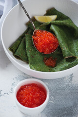 Close-up of green crepes with addition of spinach and red caviar in a white bowl, vertical shot on a white concrete surface