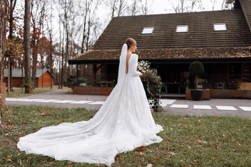 Fototapeta premium bride in a long white dress is standing in the park and holding a wedding bouquet of roses flowers and greens. young woman with hairstyle, makeup and long veil outdoors near hotel