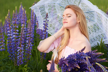 Young attractive long-haired blonde with a white lace umbrella and a bouquet of lupins among a purple field. The concept of nature and romance.