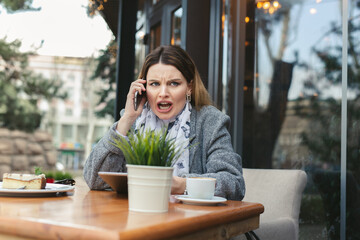 Portrait of angry young woman solving business problems shouting during phone conversation sitting in cafe terrace.Disappointed female upset about getting bad news from employee talking on cellular
