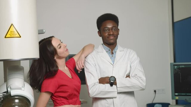 Close up portrait of team of two smiling professional doctors, African man and Caucasian woman, standing in front of ultrasound and lithotripter machines. Safety non-invasive stone treatment