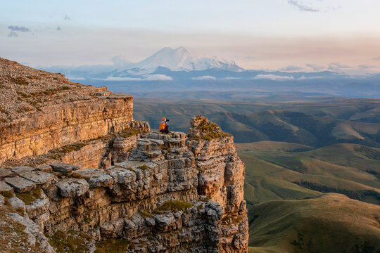 A tourist (girl) sits on the cliff edge of Bermamyt plateau against mount Elbrus and taking photos of sunset landscape. Karachay-Cherkessia, Caucasus, Russia.