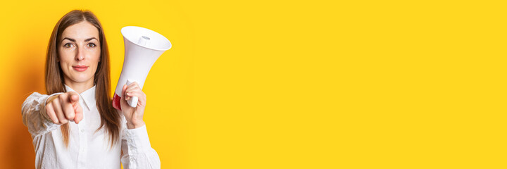 Friendly young woman holds a megaphone in her hands and points her finger at the viewer on a yellow background. Hiring concept, help wanted. Banner