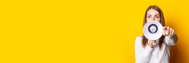Friendly young woman holds a megaphone in her hands and points her finger at the viewer on a yellow...