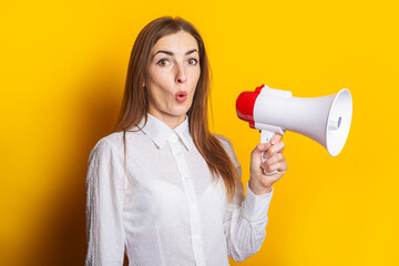 Surprised young woman in a white shirt holds a megaphone on a yellow background. Hiring concept, ad