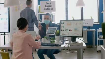 Executive manager with face mask showing statistics to her collegue using digital tablet computer. Coworkers working in new normal office keeping ing ing social distance to prevent infection with - Powered by Adobe