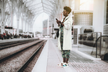 A young fancy curly-hair African woman in sunglasses, white trench, and camouflage trousers is phoning while standing on the platform of a railroad station depot and waiting for the train to travel - Powered by Adobe