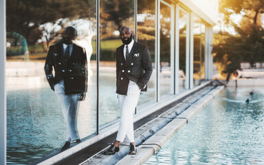 A stately mature bearded bald black guy in an elegant costume with white trousers is standing on the street, in between an artificial pond or a fountain and a glass facade, with his hand in his pocket