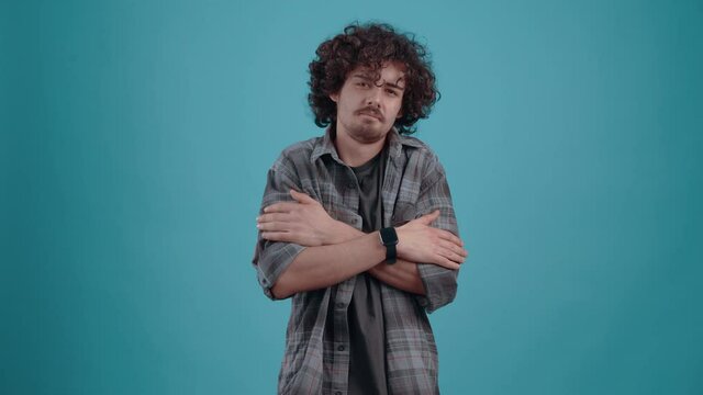 Young empathetic hipster type. Shy embraces with his hands looking puzzled to the right, finally smiling. Boy dressed in gray plaid shirt, on blue studio background