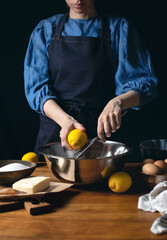 Woman in dark blue apron cooking lemon cake grating the lemon at the moment, front view
