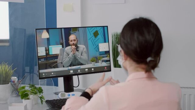 Freelancer with protection face mask talking with executive manager on computer duringvirtual meeting while sitting in new normal business office. Caucasian female working during coronavirus pandemic