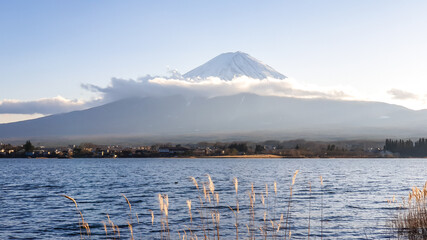 An idyllic view on Mt Fuji from the side of Kawaguchiko Lake, Japan. The magnificent mountain is surrounded by clouds. Soft reflections in the calm surface of the lake. Serenity and calmness