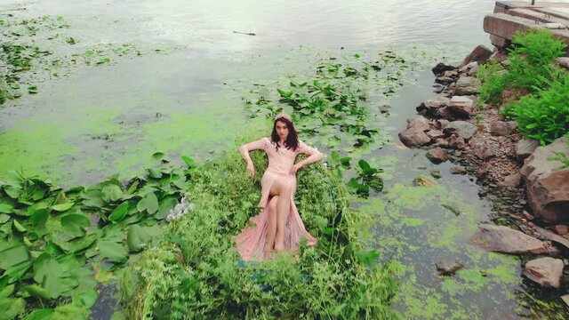 Fantasy woman fashion model sitting posing in boat throne, sexy pink peanch dress, slits on legs. Fairy Girl princess. summer nature lake green water lilies, beauty queen. gold crown. lady river nymph