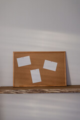 Open floating wooden shelf with a big cork board with frame and empty mock up sheets on a white wallpaper wall in a modern design living room.