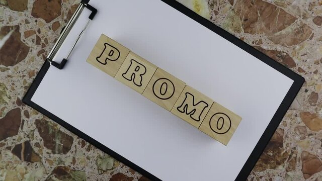 Promo concept with alphabet letters on wooden cubes arranged centered on blank white paper on a clipboard over a stone background viewed from above. Rotating table