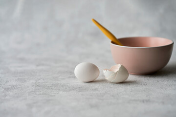 Pink bowl with a spoon on a gray textured background. Next to it is an egg and the shell of an egg. Selective focus. Copy space.