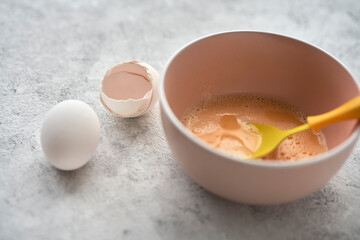 A bowl with egg mass on a gray textured background. Next to it is an egg and the shell of an egg....