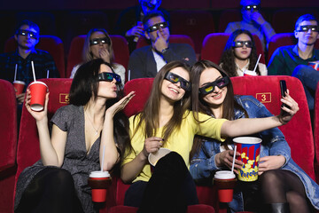 Three beautiful female friends laughing happily making a selfie together during a movie at the cinema