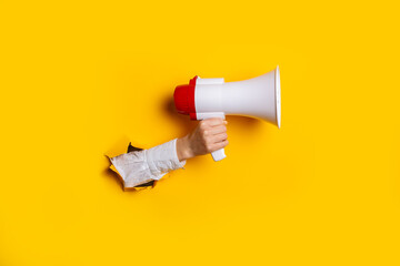 Hand holds a megaphone from a hole in the wall on a yellow background. Concept of hiring, advertising something. Banner - obrazy, fototapety, plakaty