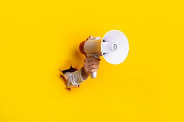 Hand holds a megaphone from a hole in the wall on a yellow background. Concept of hiring,...