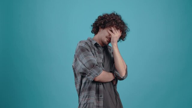 Shy, sad teenager with curly hair. With his hands over his eyes, he spies between his fingers, looking down, and finally looks into the room. Boy dressed in gray plaid shirt, on blue studio background