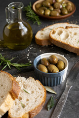 Italian sliced ciabatta bread on chopping board with herbs, extra virgin oil and marinated olives on dark grunge backdrop