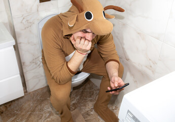 Man in cosplay costume of a cow. Guy in the funny animal pyjamas sit on the toilet and playing games on the mobile phone.
