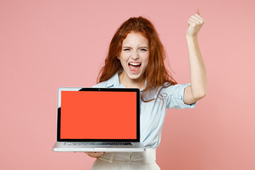 Young happy student businesswoman redhead freelancer woman 20s in blue shirt hold laptop pc computer with blank screen workspace area do winner gesture clench fist isolated on pastel pink background.