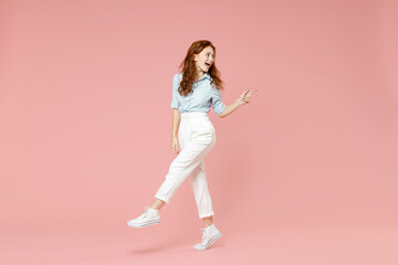 Fototapeta na wymiar Full length young student friendly smiling happy redhead woman 20s in blue shirt pants walk point index finger back aside on workspace area mock up isolated on pastel pink background studio portrait.