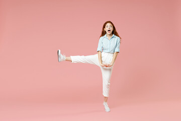 Full length body of young student positive smiling happy caucasian redhead woman 20s wear blue shirt pants standing with outstretched raised up leg isolated on pastel pink background studio portrait.