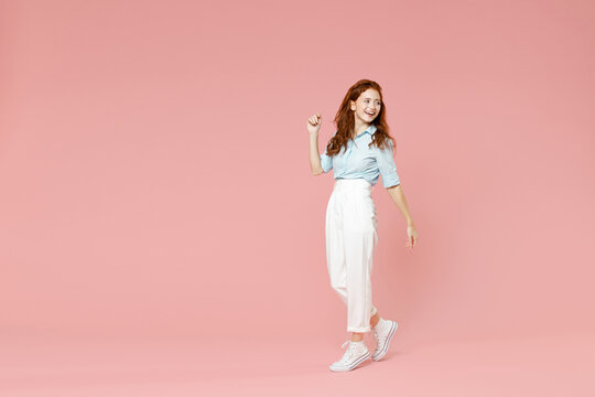 Full Length Body Of Young Student Positive Smiling Happy Caucasian Redhead Woman 20s Wearing Blue Shirt Pants Walking Going Looking Back Aside Isolated On Pastel Pink Color Background Studio Portrait.