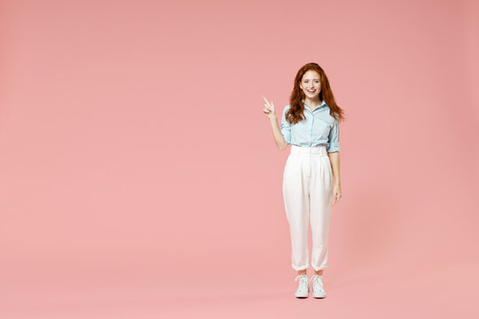 Full length body young student friendly smiling happy redhead woman 20s in blue shirt pants walk point index finger aside on workspace area mock up isolated on pastel pink background studio portrait.