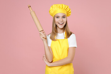 Teen happy girl chef cook confectioner baker housewife mother's helper in yellow apron white t-shirt toque cap hold rolling pin isolated on pastel pink background studio portrait. Food cake concept.