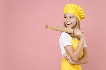 Teen side view girl chef cook confectioner baker housewife mother's helper wear yellow apron white t-shirt cap hold on shoulder rolling pin spread hand isolated on pastel pink background Food concept.
