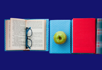 Open textbook, pile of books in colorful covers and apple on blue background. Back to school distance home education. .