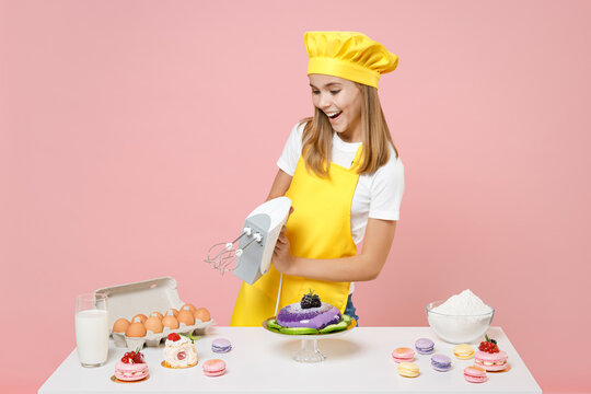 Teen girl chef cook confectioner baker in yellow apron toque cap at table use kitchen mixer do winner gesture clench fist isolated on pink background studio Mousse cake food process workshop concept