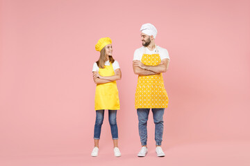 Full length body of teen girl dad man father's helper chef cook confectioner baker wear yellow apron toque cap hold hands crossed folded look to each other isolated on pink background studio portrait.